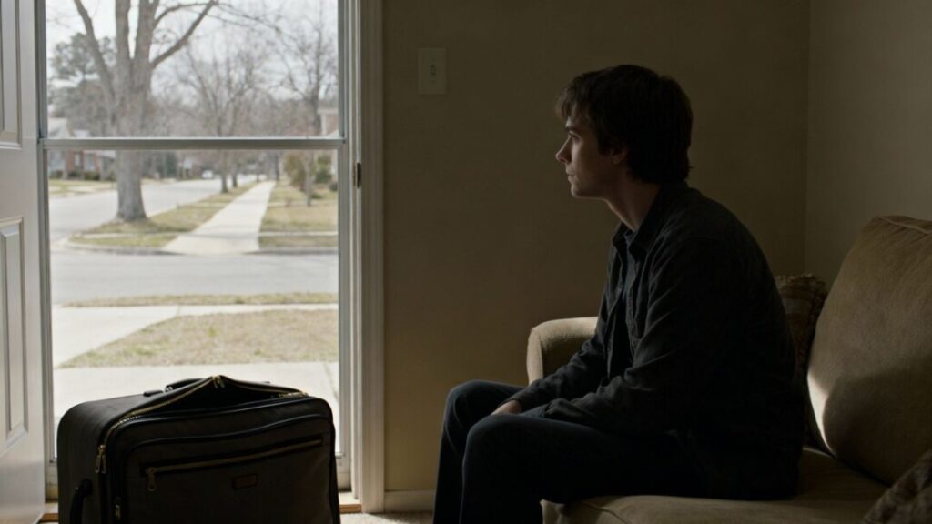 Person looking out window, suitcase by door, quiet street.